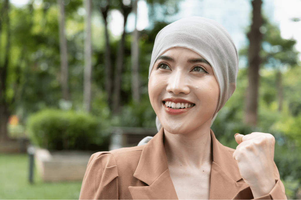 Smiling woman wearing a headscarf and brown blazer raising her fist in a gesture of triumph, standing outdoors in a green park.